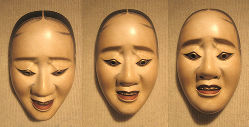 Three masks are arranged side by side. The masks are almost identical, but with slightly different facial expressions resulting from the masks being at different angles. The first mask is tilted downward and has downcast eyes. The second mask is shown straight on and is directing its gaze slightly higher than the first. The third mask is tilted upwards so its gaze is directed more upward.