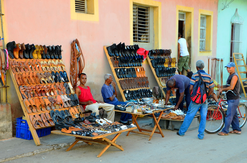 Some tables filled with shoes available for sale on the street. A few people peruse the shoes.