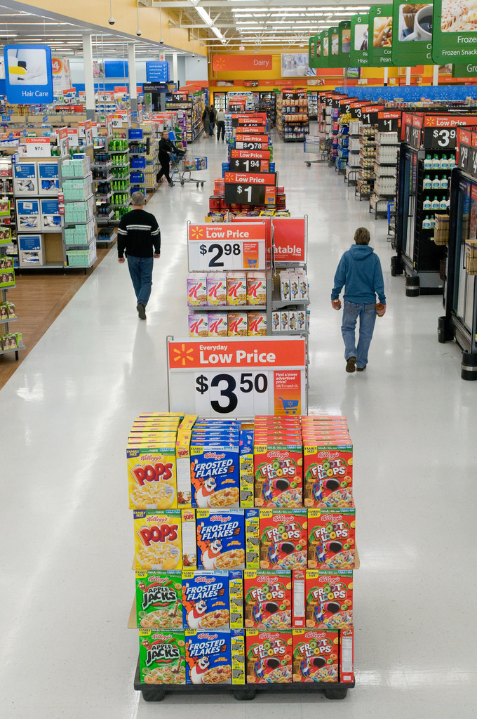 Photo of a shopping aisle in Walmart. Large center-aisle displays of breakfast cereal are in the foreground. Three shoppers are in the background.