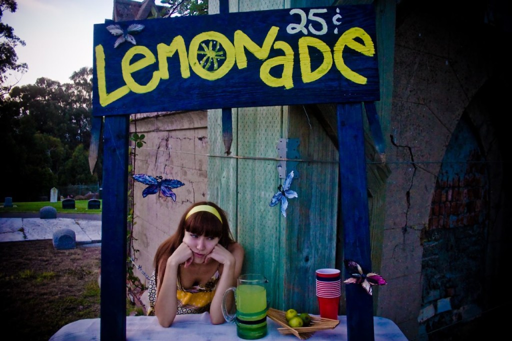 Photo of a colorful lemonade stand set up in a cemetery. The discourage young woman running the stand is shown with her head in her hands.