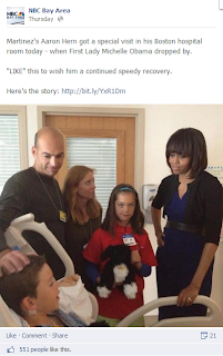Michelle Obama visits a child and the child's family in a hospital. The child is in bed; his family stands next to him, along with Michelle Obama. 