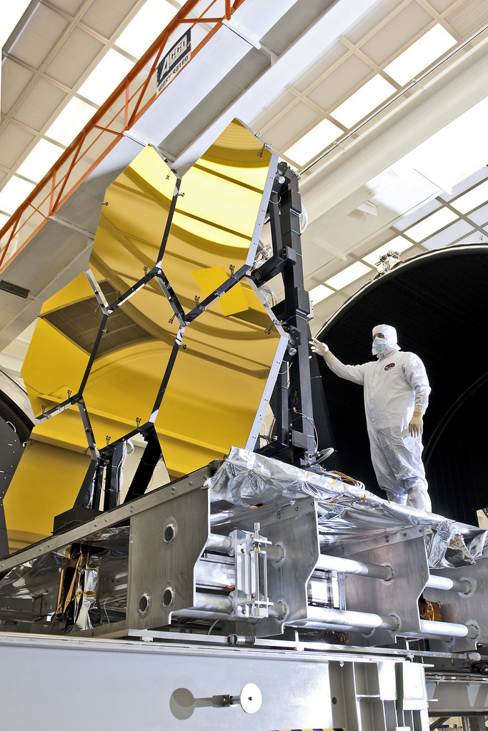 Photo inside NASA space flight center. Man in a white protective suit is holding on to part of the structure that contains six mirror segments for the James Webb Space Telescope.