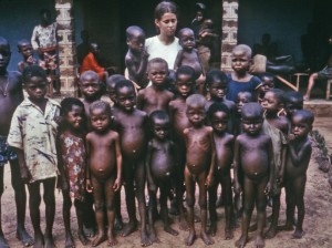 White woman standing with malnutritioned African children, many who display kwashiorkor, or the swollen bellies.