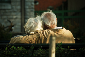 Elder couple sitting and cuddling on a park bench. Image is taken from behind them.