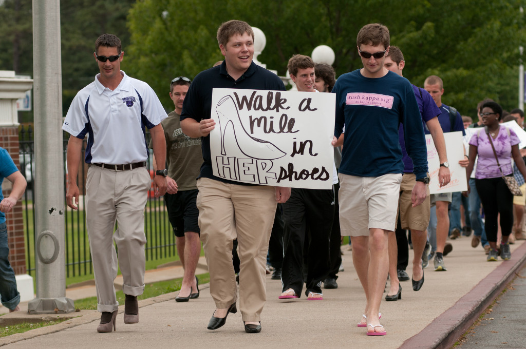 A group of male students is walking; most are wearing women's shoes, and one holds a sign that reads,