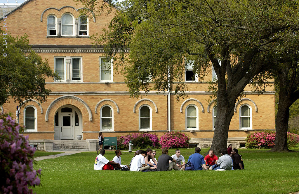 A group of students sits in a circle on the lawn in front of a college building.