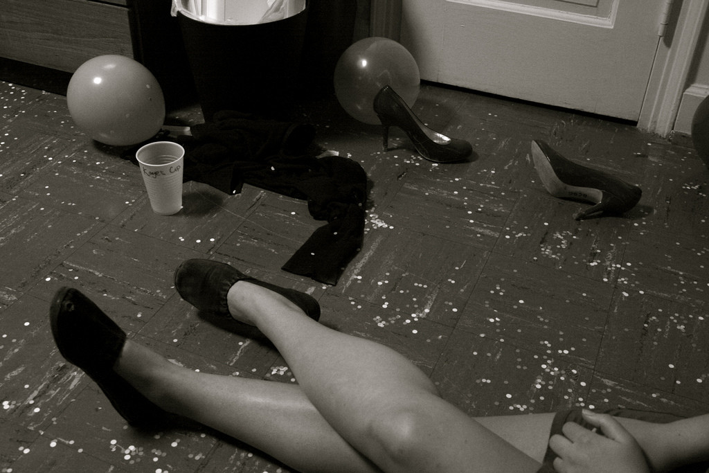 Photo of a woman's legs. Though mostly out of the frame, she appears to be sitting on the floor at a party. Next to her, on the floor, is a pair of high-heeled women's shoes, a party balloon, a plastic beer cup.