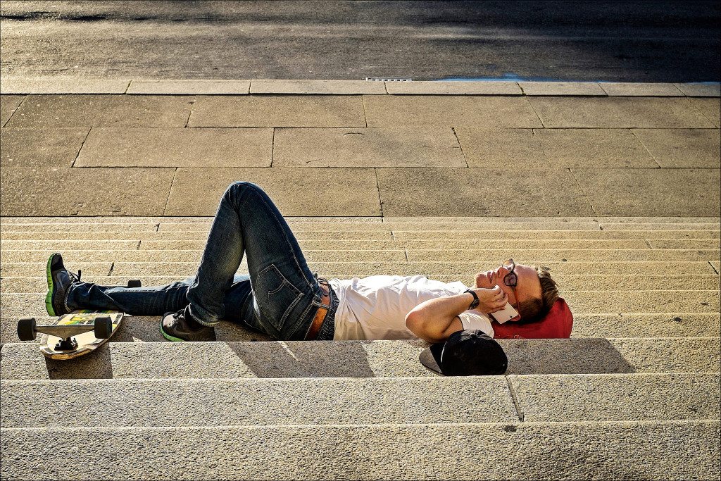 Man lies comfortably on his back in the sun on public steps, his skateboard at this feet, phone in hand.