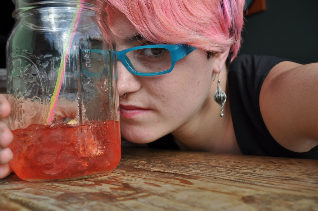 Young female college student with short pink hair and blue glasses is frowning and leaning over a table. In her hand is a jarful of pink punch or juice with a straw.