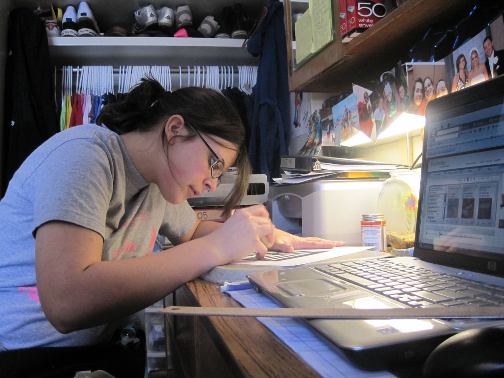 Photo of a young woman focused intently on writing something. She is leaned over her desk; her keyboard and computer are beside her.