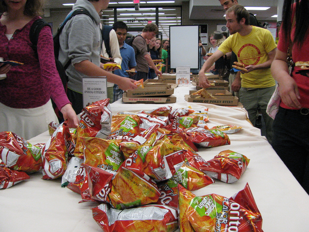 Photo of a college cafeteria. Students push lunch trays past a pile of Doritos and pizza.