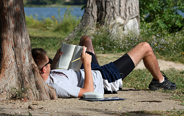 Photo of a man lying on the ground, against a tree, holding a book and a pencil in hand