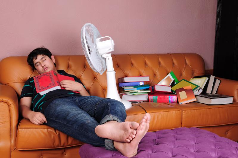 Photo of a male student asleep on a couch amid a pile of book. A fan is aimed at his face, and a book lies open on his chest
