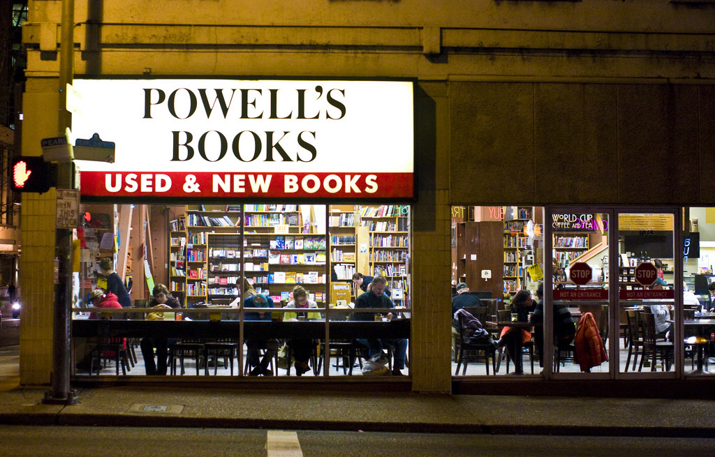 A lit-up storefront for Powell's Books bookstore. Large windows show tall shelves full of books and tables where people are sitting, eating, and reading.