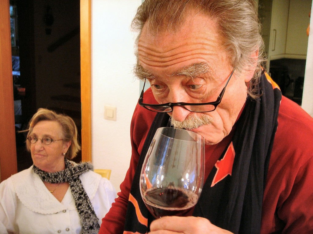 Picture of an elderly man sniffing a glass of red wine. A woman is in the background.