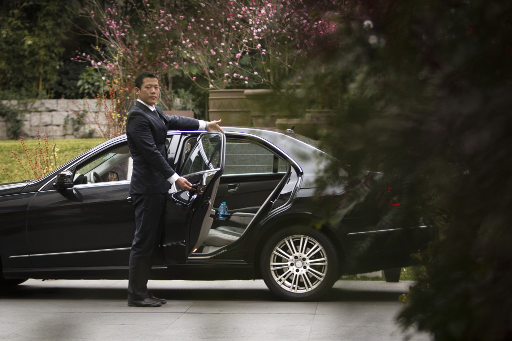 An Uber driver dressed in a suit stands beside a an elegant black sedan, opening the back door. Shanghai, China.