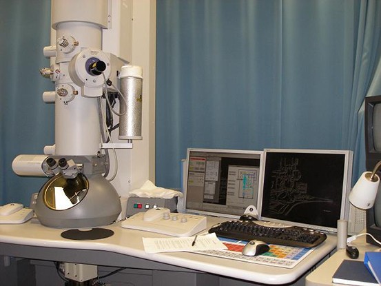 Photograph A shows a transmission electron microscope: a large-tube shaped machine attached to a desk next to a computer. Photograph b shows a scanning electron microscope: a machine with many projections sitting on a desk next to a computer.