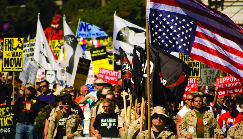 An image of a group of people, several of whom are holding flags and signs. One of the signs reads 