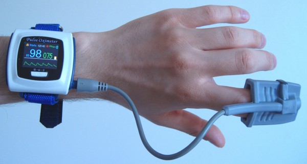 A person is measuring the amount of oxygen in blood and metabolic rate using a pulse oxymeter. The pulse oxymeter is strapped to the person’s wrist, and the index finger is inside the clip.