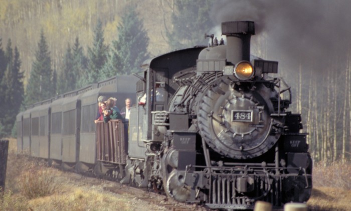 A steam engine and several passenger cars are shown traveling down a train track. The train has some people on board.