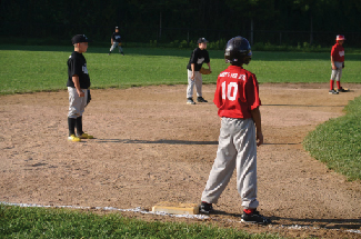 A photograph of children playing baseball is shown. Five children are in the picture, two on one team, and three on the other.