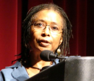 Woman wearing glasses, speaking into a microphone on a lectern