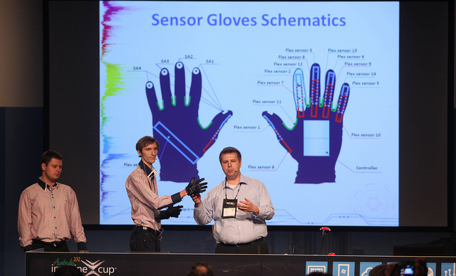 One man wears a sensor glove while another man points at the glove and speaks into a microphone. Behind them is a large powerpoint slide showing schematics for the sensor glove.