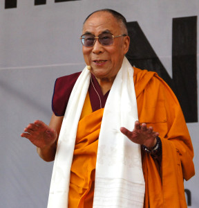 Dalai Lama wearing a white sash and saffron robes, hands outstretched facing down