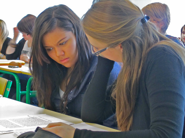 Photo of two young women reading the same sheet of paper on a desk, in a crowded classroom
