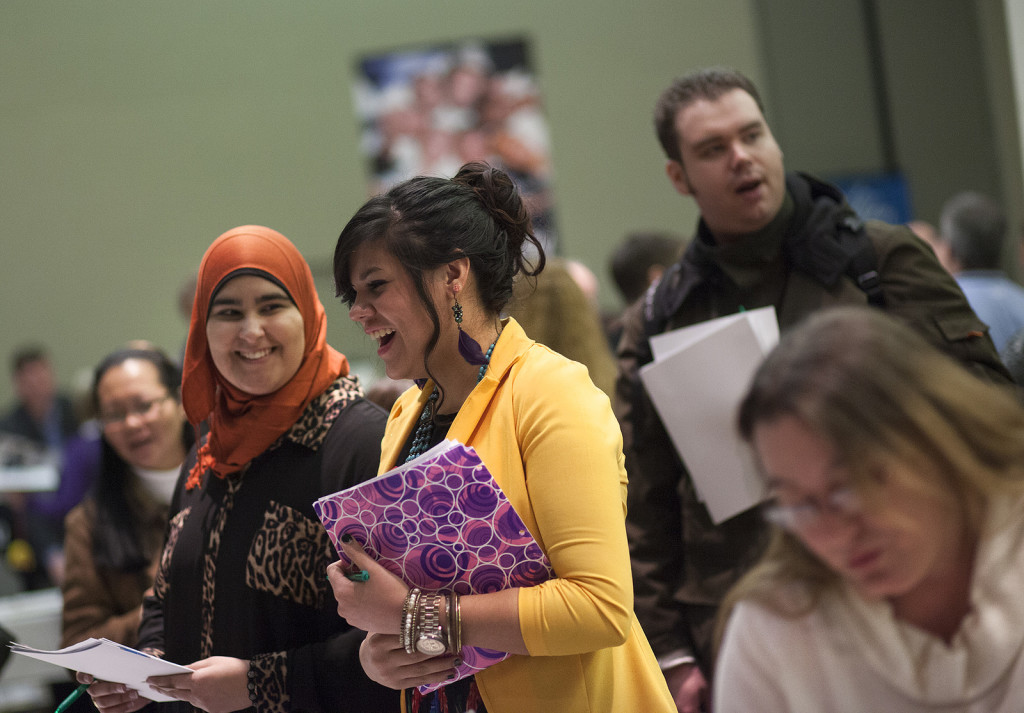 Photo of room full of active people. Central are two women, one wearing a head scarf, holding notebooks and laughing.