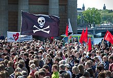 Protestors standing in a crowded street holding red flags and a large Jolly Rogers flag.