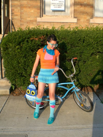 A young woman in brightly colored clothes and carrying an owl handbag is shown standing in front of a vintage blue bicycle, a large hedge, and a town house.