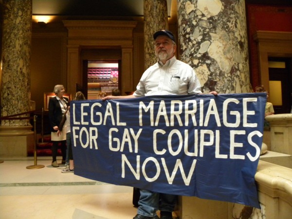 An older man with a gray beard, wearing a baseball cap, buttoned-up shirt, and jeans, is shown in a marbled lobby with columns holding a blue banner reading “Legal Marriage for Gay Couples Now.”