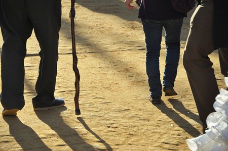 The legs of three men, one using a cane, are shown from behind walking on a dirt surface.
