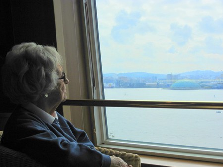 An older woman with white hair and glasses is shown looking out a window, across a body of water.