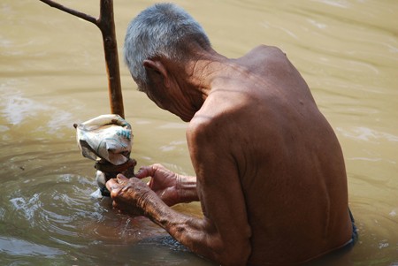 A shirtless elderly man is shown manipulating a large tree branch while standing waist-deep in a river.