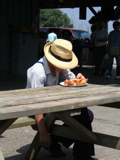 A young Mennonite boy in a straw hat is shown eating a piece of pizza.