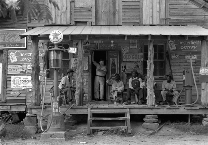 Black-and-white photo of a country store on a dirt road (Gordonton, North Carolina, 1939), showing four African American men sitting in chairs on the porch and a white man (brother of the store owner) standing in the doorway.