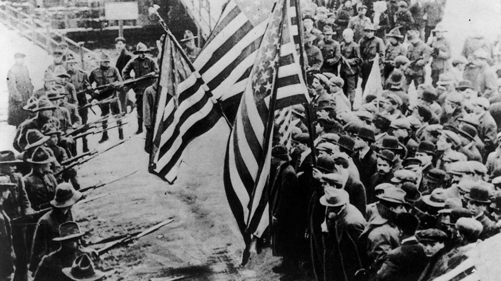 A group of people holding large American flags face off with a smaller group of people holding bayonets.