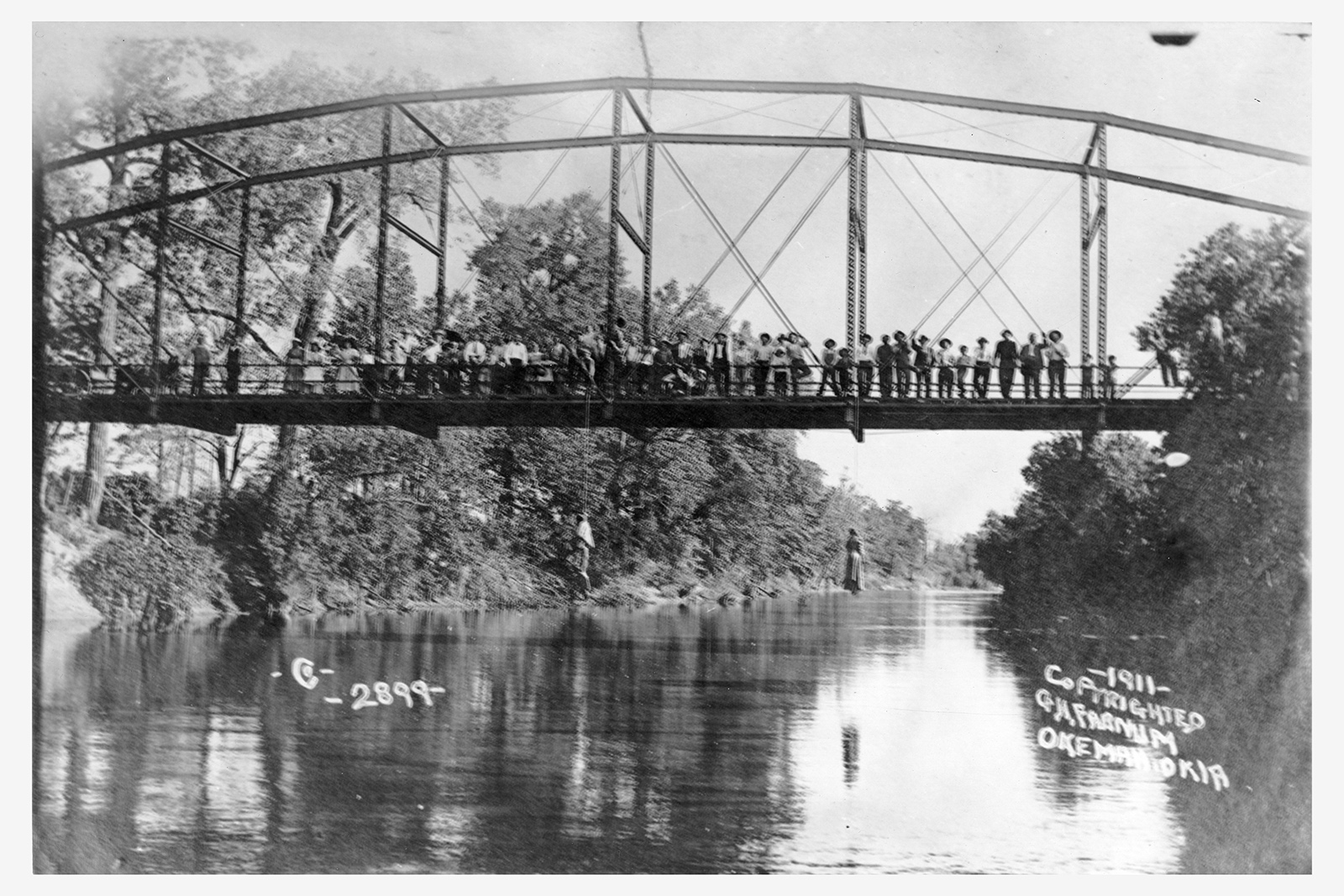 A photograph of a crowd of people standing on a bridge, from which two dead black people are dangling.