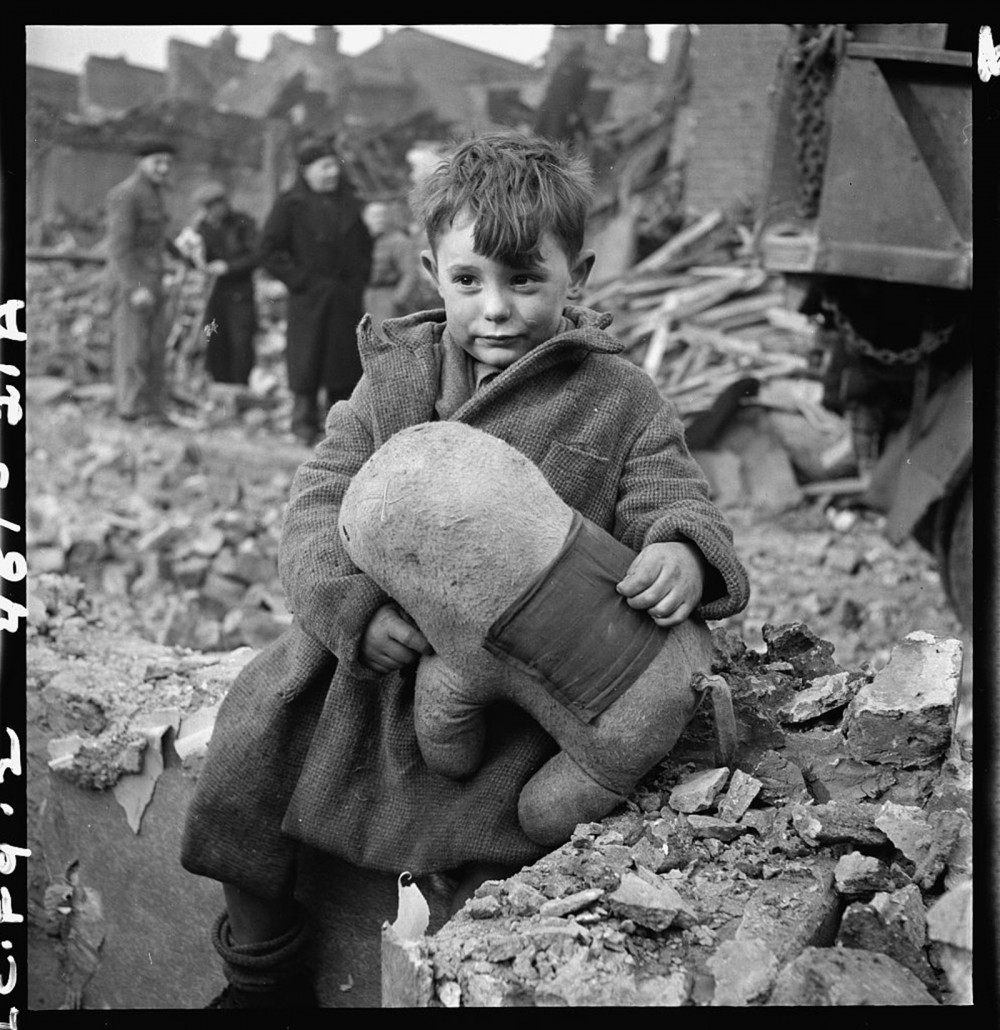 A small boy holding a stuffed toy in the midst of rubble.