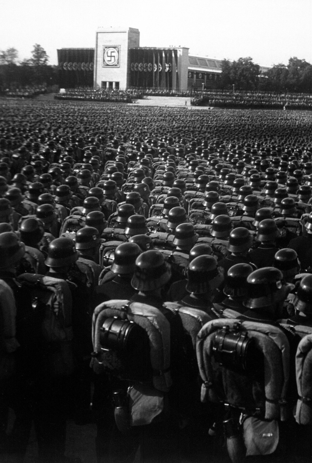 Thousands of soldiers in formation beneath a sign bearing the Nazi symbol.