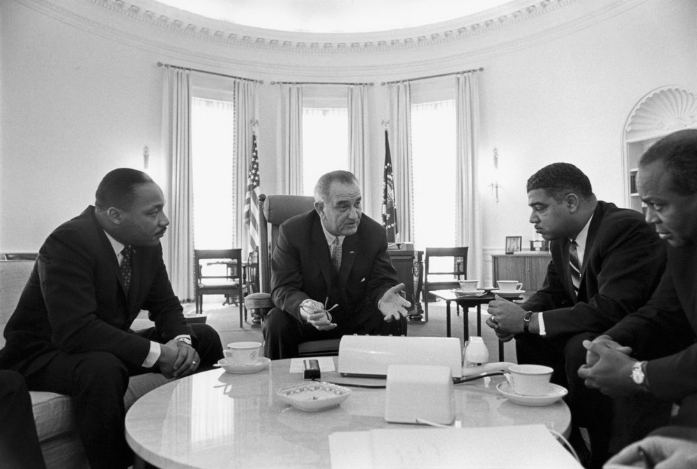 LBJ sits in a chair at the center of a table with Martin Luther King, Jr. and other Civil Rights leaders.