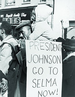 A photograph shows a group of African Americans marching on the street in Selma, Alabama. In the foreground, a man with a small child on his shoulders carries a sign that reads “President Johnson/Go to Selma now!”