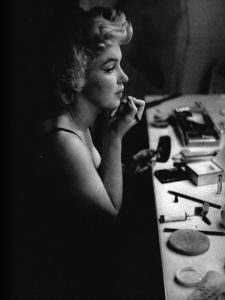 Black and white photo of Marilyn Monroe applying lipstick in a dressing room mirror