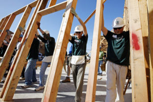 Men in hard hats and volunteer tee shirts raising a wall frame for a house