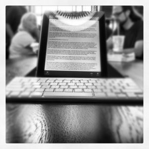Black and white photo of a tablet displaying text and wireless keyboard, on a table in a cafe with people in the background