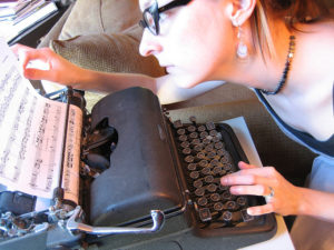 Woman peering at a page in the typewriter before her, which contains musical scores.