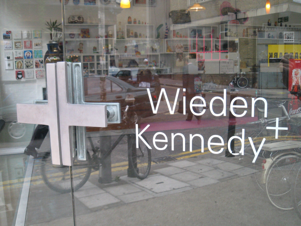 Photo of glass front door to Wieden + Kennedy building. Small shelves filled with objects are visible through the door; so is the reflection of the sidewalk and street out front—a bike and parking meter are visible.