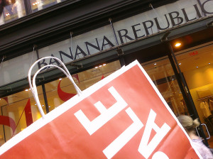 Photo of Banana Republic storefront. In foreground, partial view of a large red shopping bag, with the word SALE printed in white.